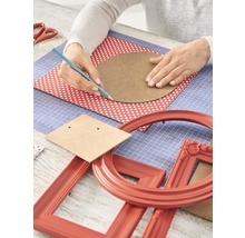 Basteltisch mit Händen, die einen Kreis auf ein Blatt Papier zeichnen, umgeben von Bastelmaterialien und Bilderrahmen