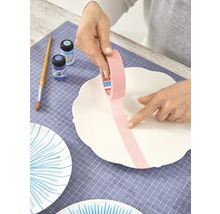Eine Person klebt Malerkrepp auf eine weiße Keramikplatte, um sie zu dekorieren. Pinsel, Farben und weitere bemalte Platten sind sichtbar.