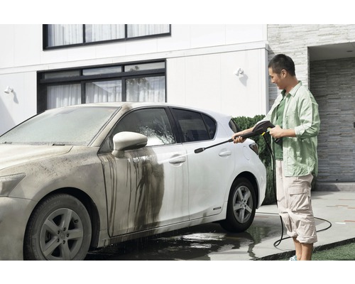 Mann reinigt ein schmutziges Auto mit einem Hochdruckreiniger vor einem Wohnhaus.