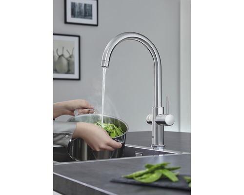 Küchenszene mit Spültischarmatur, an der eine Person einen Topf mit Gemüse befüllt