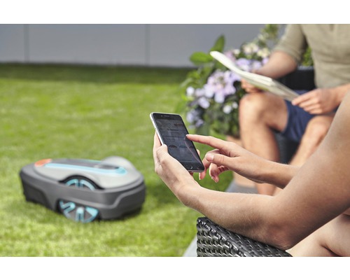Person bedient Rasenmähroboter mit Smartphone im Garten.