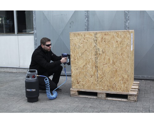 Ein Mann benutzt einen Druckluftnagler an einer Holzkiste, die auf einer Palette steht. Ein Kompressor der Marke Aerotec ist ebenfalls zu sehen.