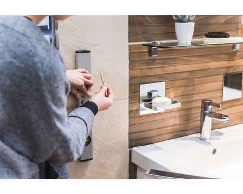 Eine Person verwendet eine Wasserwaage und einen Bleistift im Badezimmer, um eine gerade Linie an der Wand zu markieren. Im Hintergrund sind ein Waschbecken, eine Armatur und ein Seifenspender zu sehen.