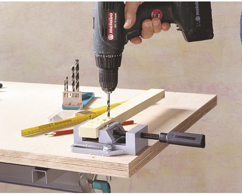 Eine Person bohrt mit einem Akkubohrer ein Holzstück ein, das mit einem Schraubstock fixiert ist.