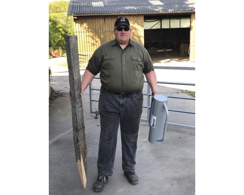 Mann hält Holzpfosten und Zaunrüttler vor einem Stallgebäude