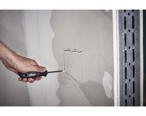 Eine Person schneidet mit einem Cuttermesser ein Loch in eine Gipskartonwand.