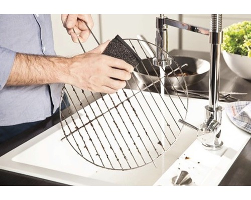 Eine Person reinigt einen Grillrost mit einem Schwamm über einem Spülbecken.