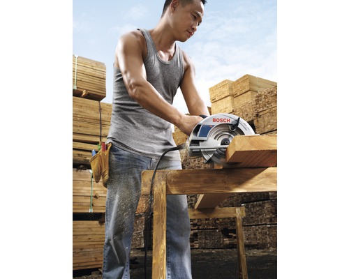 Ein Mann schneidet mit einer Kreissäge ein Holzbrett, im Hintergrund Holzstapel.