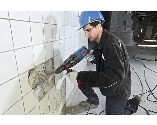 Handwerker mit Schutzbrille, Gehörschutz und Schutzhelm arbeitet mit einem Bohrhammer an einer gefliesten Wand