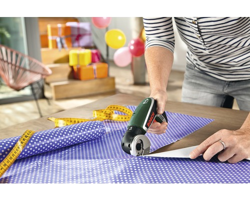 Szene mit einer Person, die mit einem Akku-Cutter Geschenkpapier zuschneidet, zusammen mit einem Maßband und Geschenkverpackungen