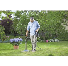Mann bedient eine Tiefbrunnenpumpe im Garten, umgeben von Blumen und Grünflächen.