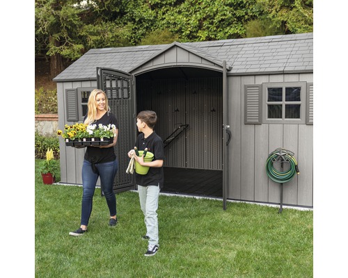 Gartenszene mit einem Gerätehaus, einer Frau und einem Jungen, die Gartenartikel tragen