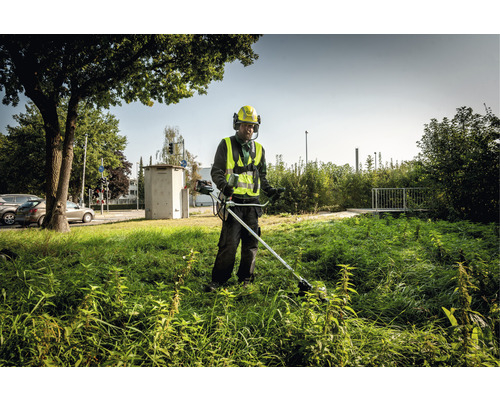 Mann mäht mit einem Rasentrimmer hohes Gras in einer städtischen Umgebung.