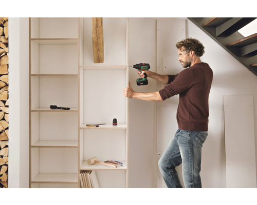 Mann montiert mit einem Akkuschrauber ein Regal im Raum, Werkzeuge liegen bereit.