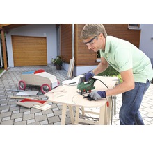Mann arbeitet mit einer Stichsäge an einem Holzbrett im Freien, im Hintergrund ein Holzauto