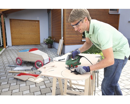 Mann arbeitet mit einer Stichsäge an einem Holzbrett im Freien, im Hintergrund ein Holzauto