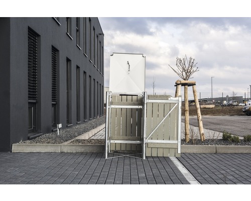 Mülltonnenbox mit Holzverkleidung und Metalldeckel auf einem gepflasterten Platz vor einem Gebäude.