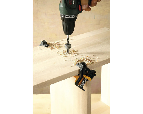 Eine Bohrmaschine bohrt in ein Holzbrett, befestigt mit einem Winkelspanner.
