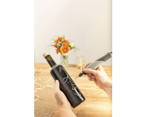 Eine Person graviert mit einem Dremel Graviergerät eine Glasflasche mit Initialen und einem Datum.