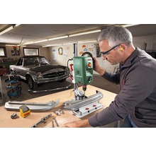 Werkstattszenario mit einer Bosch Tischbohrmaschine beim Bohren eines Werkstücks, ein Mann trägt dabei Schutzbrille und Gehörschutz.