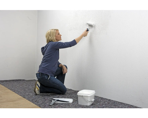 Frau streicht Wand mit Farbroller in hellem Raum. Eimer, Abstreifgitter und Abdeckvlies sind ebenfalls vorhanden.