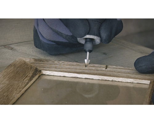 Eine Person mit Handschuhen verwendet ein Multifunktionswerkzeug an einem Holzrahmen.