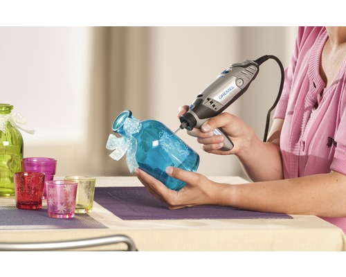 Frau graviert ein blaues Glas mit einem Dremel Multifunktionswerkzeug. Dekorative Glaswaren im Hintergrund.