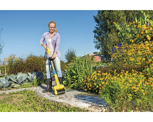 Frau entfernt Unkraut mit einer elektrischen Fugenbürste auf einem Gartenweg.