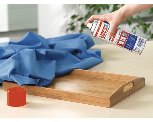 Eine Hand sprüht Tesa Kontaktspray auf ein blaues Tuch, das auf einem Holztablett liegt