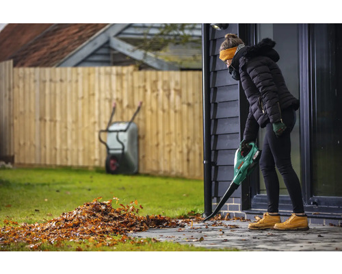 Eine Frau benutzt einen Laubbläser, um Laub auf einer Terrasse zu entfernen. Im Hintergrund sind ein Zaun und eine Schubkarre zu sehen.