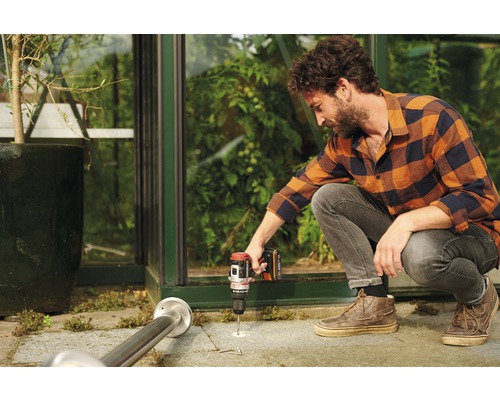 Ein Mann bohrt mit einem Akkuschrauber ein Loch in den Boden vor einem Gewächshaus.