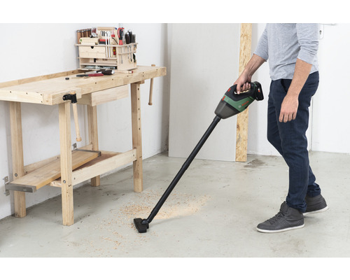 Mann saugt Holzspäne mit einem Akku-Staubsauger neben einer Werkbank auf.