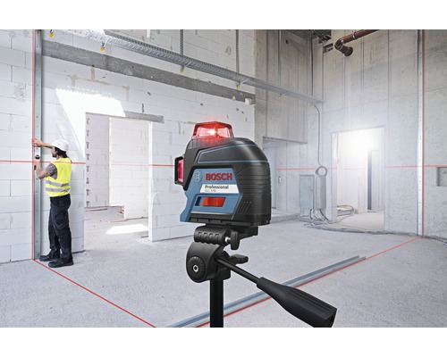 Bosch Logo. Baustelle mit Kreuzlinienlaser auf Stativ, der rote Laserlinien an Wänden und Boden projiziert. Arbeiter mit Schutzhelm und Warnweste richtet eine Metallschiene aus.