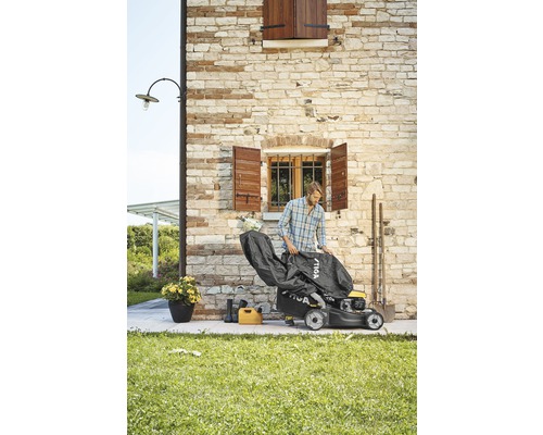 Mann deckt Rasenmäher mit Schutzhülle ab. Im Hintergrund befindet sich eine Hauswand.