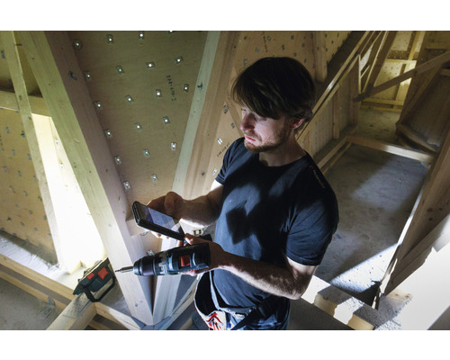 Mann fotografiert mit einem Mobiltelefon eine Bohrmaschine vor einem Holzdachstuhl