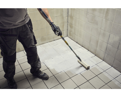 Handwerker streicht Fliesenboden mit Farbroller in einem Badezimmer.
