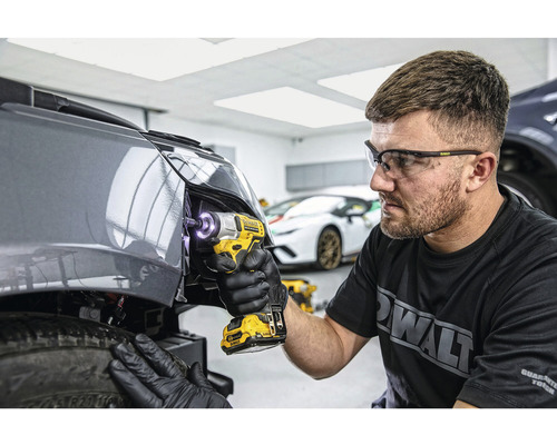 Ein Mann befestigt mit einem Akku-Schlagschrauber ein Autoteil. Er trägt eine Schutzbrille und Handschuhe. Im Hintergrund sind weitere Werkzeuge und Autos zu sehen.