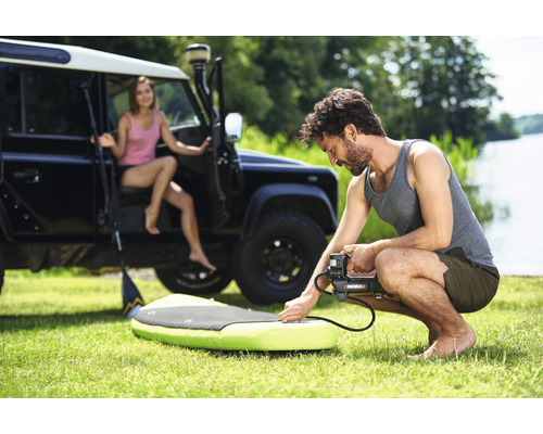 Mann pumpt Stand-Up-Paddleboard mit einer elektrischen Pumpe auf, im Hintergrund eine Frau und ein Geländewagen.