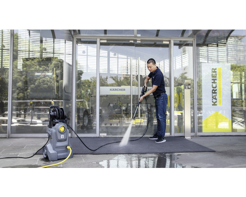 Ein Mann benutzt einen Hochdruckreiniger vor einem Gebäude mit Glasfassade. Der Hochdruckreiniger steht auf dem Boden und ist mit einem gelben Stromkabel verbunden.