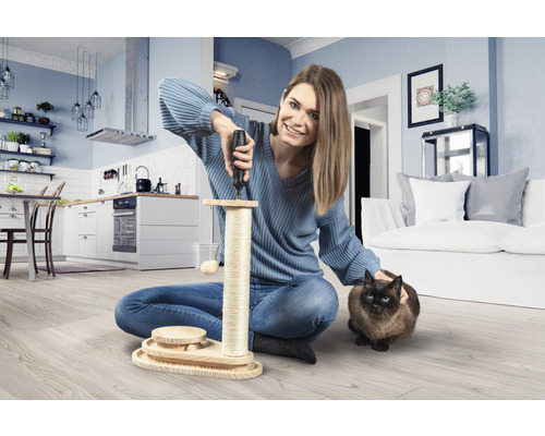 Frau montiert einen Katzenkratzbaum mit einem Schraubenzieher in einem Wohnzimmer. Eine Katze sitzt daneben.