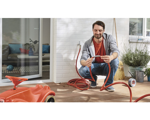 Mann schließt einen Gartenschlauch an einen Wasserhahn an, im Hintergrund ein Haus mit Terrasse