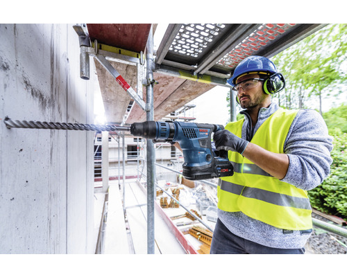Ein Mann bohrt mit einem Bohrhammer ein Loch in eine Betonwand auf einem Baugerüst.