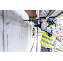 Ein Bauarbeiter bohrt mit einem Bohrhammer ein Loch in eine Betonwand auf einem Gerüst.