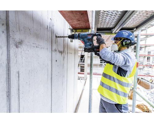 Ein Bauarbeiter bohrt mit einem Bohrhammer ein Loch in eine Betonwand auf einem Gerüst.