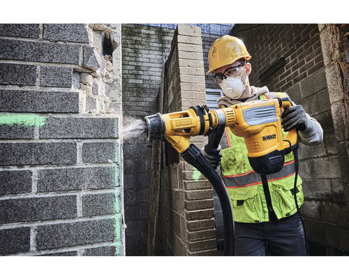 Ein Bauarbeiter in Schutzausrüstung benutzt einen Abbruchhammer zum Bearbeiten einer Ziegelwand. Das Gerät ist mit einem Staubabsaugungsaufsatz versehen. DeWalt Logo.