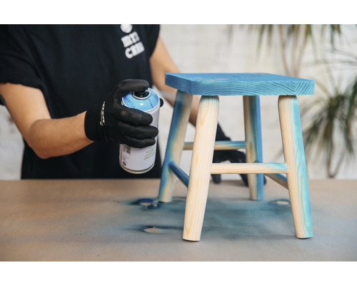 Eine Person sprüht eine blaue Farbe auf einen kleinen Holzschemel.