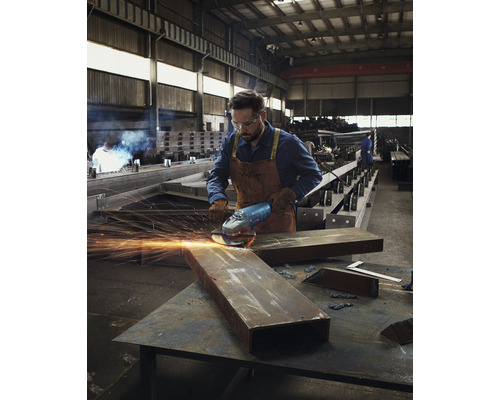 Ein Mann in Arbeitskleidung benutzt in einer Werkstatt einen Winkelschleifer an einem Metallstück.