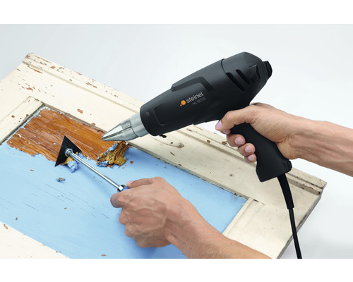 Person entfernt Farbe von einem Fensterrahmen mit einer Heißluftpistole und einem Farbschaber