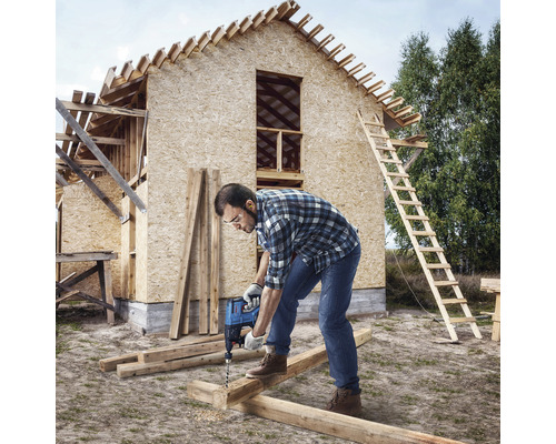 Handwerker bohrt mit einer Bohrmaschine ein Holzbrett vor dem Rohbau eines Hauses.