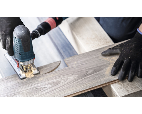 Eine Stichsäge schneidet ein Laminatbrett. Schutzhandschuhe werden getragen.